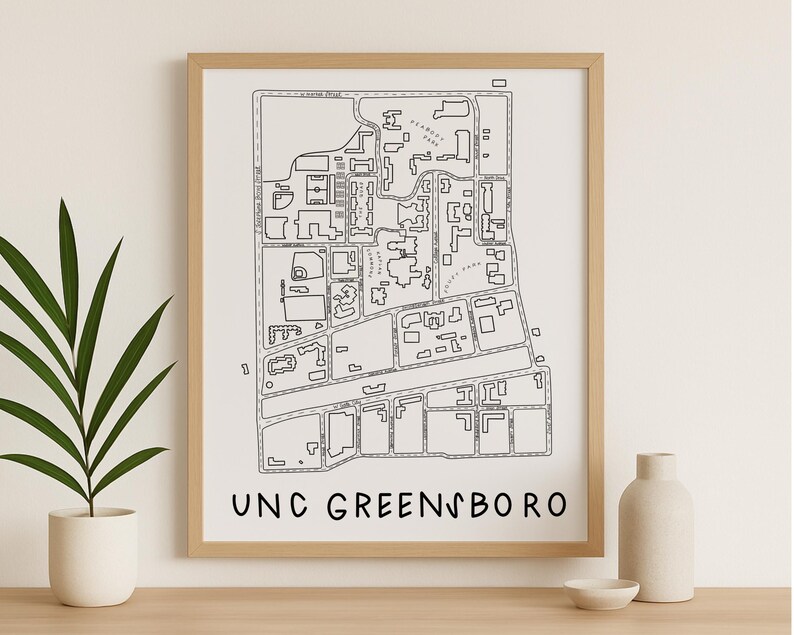 Op de afbeelding: Een ingelijste zwart-wit kaartafdruk van UNC Greensboro. De kaart toont de campusindeling met straten, gebouwen en groene ruimtes. De woorden "UNC GREENSBORO" zijn gedrukt in een vetgedrukte, schreefloze letter onderaan de afdruk. De lijst heeft een licht houten kleur.