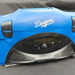 Könnte beinhalten: Blauer und schwarzer tragbarer Radiokühler mit dem Wort "Dodgers" in weißen Buchstaben auf der Oberseite. Das Radio hat ein schwarzes Gesicht mit Drehknöpfen für die Abstimmung und Lautstärke.