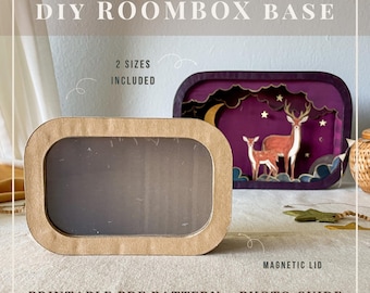 May include: A DIY roombox base kit featuring a rectangular cardboard frame with a clear window and a magnetic lid. The text reads DIY ROOMBOX BASE, 2 SIZES INCLUDED, MAGNETIC LID, and PRINTABLE PDF PATTERN + PHOTO GUIDE. A finished purple diorama with deer is visible.