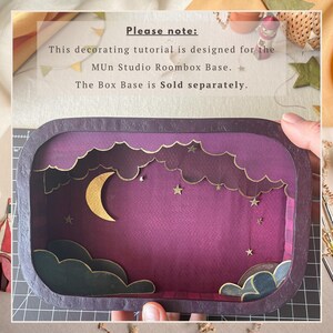 May include: A rectangular shadow box frame featuring a layered paper craft night sky scene. The design includes a gold crescent moon, small gold stars, and dark purple clouds against a deep purple background, all enclosed in a dark purple frame.