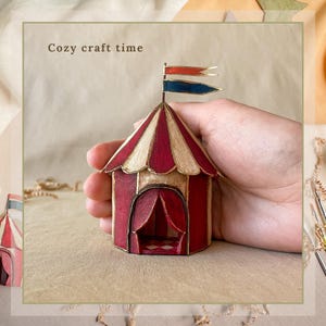 May include: A miniature circus tent with red and cream stripes, a red door, and two small flags. The text "Cozy craft time" is above the tent. The tent is held in a hand.
