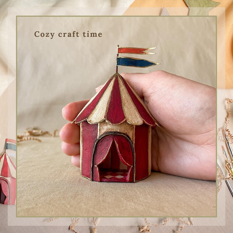 May include: A miniature circus tent with red and cream stripes, a red door, and two small flags. The text "Cozy craft time" is above the tent. The tent is held in a hand.