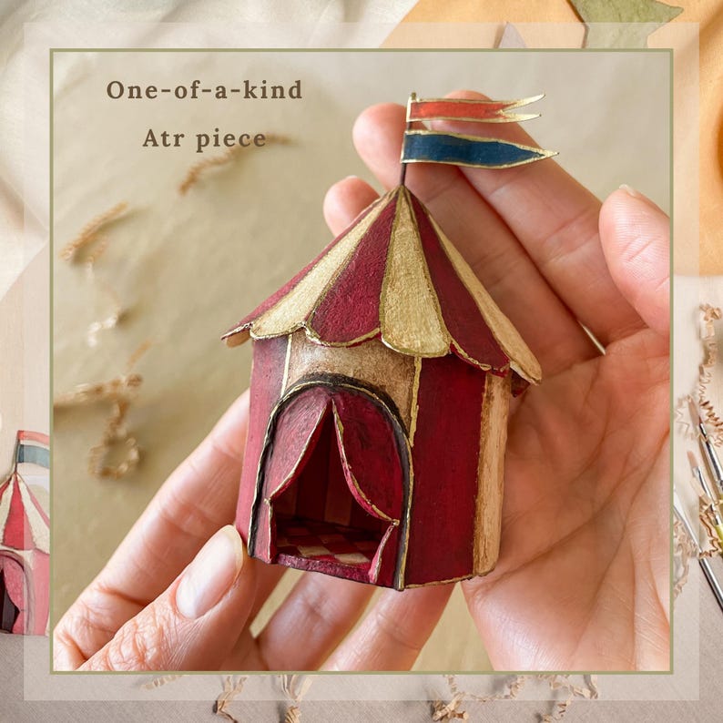 May include: A miniature circus tent with a red and gold striped roof and a red curtained entrance. The tent has a small flag on top with red, blue, and gold accents. The text "One-of-a-kind Atr piece" is visible.