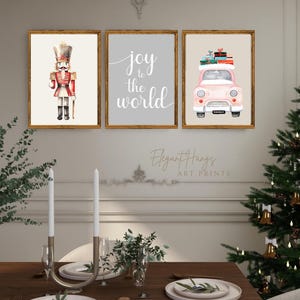Könnte beinhalten: Drei gerahmte Kunstdrucke mit Weihnachtsmotiv. Einer zeigt einen Aquarell-Nussknacker, ein anderer die Worte „joy to the world“ und der dritte ein rosa Auto mit Geschenken. Die Rahmen sind goldfarben und die Kunstwerke sind über einem Esstisch ausgestellt.