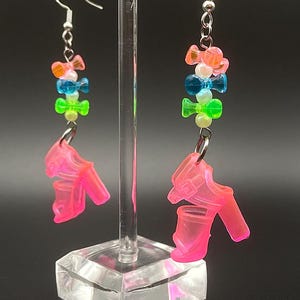 May include: A pair of neon pink high heel earrings with silver hooks. The earrings feature a colorful beaded design with orange, blue, and green beads. The earrings are displayed on a clear acrylic stand against a dark background.