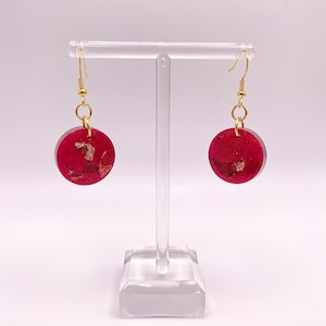 May include: A pair of red circle earrings with gold flecks. The earrings are on a clear display stand.