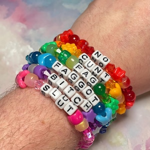 May include: A stack of colorful beaded bracelets spelling out offensive words. The bracelets feature a variety of bead shapes and colors, including red, orange, yellow, green, blue, purple, and pink. The bracelets are worn on a wrist.