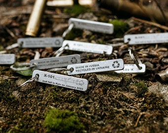 May include: A collection of small, silver metal keychains with various inscriptions, including "Made in Russia Recycled in Ukraine" and "X-101", scattered on a bed of green moss and brown dirt. Some spent bullet casings are also visible.