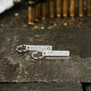 May include: Two silver keychains with the text "MADE IN RUSSIA RECYCLED IN UKRAINE" and "X-101" are displayed on a dark, textured surface. The keychains have a circular ring. In the background are several brass-colored bullet casings.