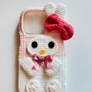 Puede incluir: Una funda para tel&eacute;fono de crochet rosa y blanco con un conejito de dibujos animados con un lazo rosa. La funda tiene una ventana de pl&aacute;stico transparente para la lente de la c&aacute;mara.