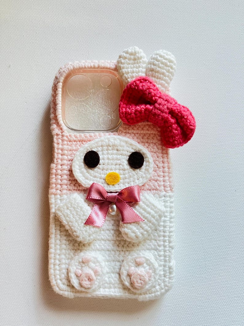 Puede incluir: Una funda para tel&eacute;fono de crochet rosa y blanco con un conejito de dibujos animados con un lazo rosa. La funda tiene una ventana de pl&aacute;stico transparente para la lente de la c&aacute;mara.