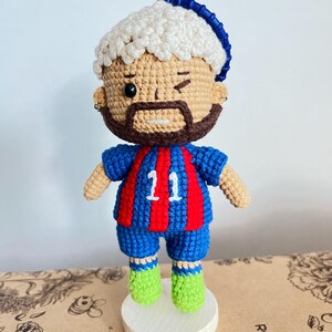 May include: A crocheted doll featuring a figure with light brown skin, a dark brown beard, and white curly hair topped with a blue headband. The doll wears a blue and red striped jersey with the number 11 in white, blue shorts, and bright green shoes.