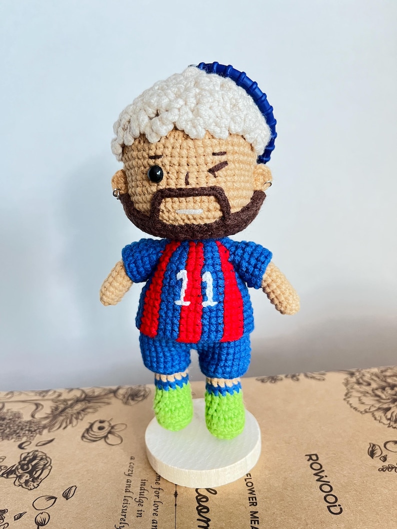 May include: A crocheted doll featuring a figure with light brown skin, a dark brown beard, and white curly hair topped with a blue headband. The doll wears a blue and red striped jersey with the number 11 in white, blue shorts, and bright green shoes.
