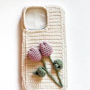 Puede incluir: Una funda de tel&eacute;fono de ganchillo con un dise&ntilde;o floral. La funda es beige con una flor rosa y hojas verdes.