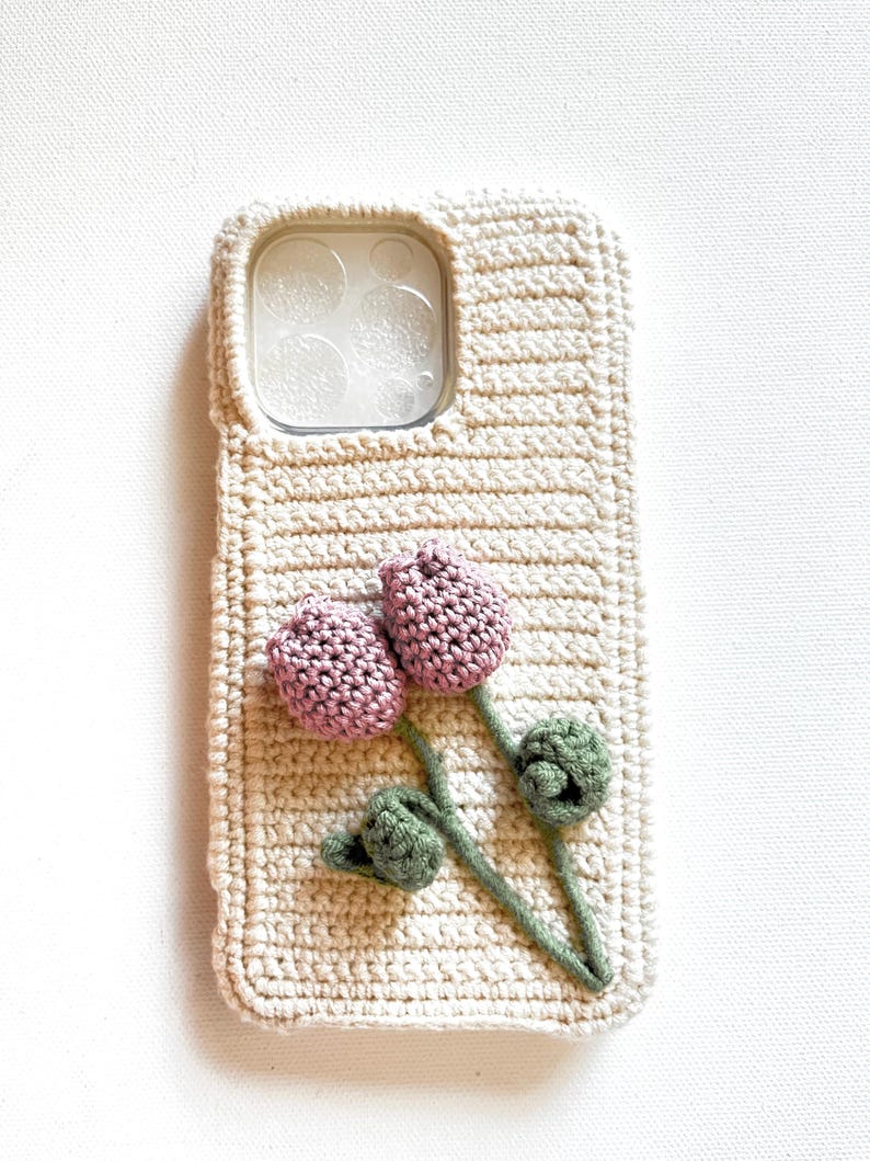 Puede incluir: Una funda de tel&eacute;fono de ganchillo con un dise&ntilde;o floral. La funda es beige con una flor rosa y hojas verdes.