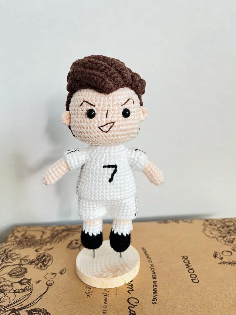 K&ouml;nnte beinhalten: Eine handgefertigte H&auml;kelpuppe, die eine Figur mit braunen Haaren darstellt, bekleidet mit einem wei&szlig;en Trikot mit der aufgestickten schwarzen Nummer 7 auf der Brust, wei&szlig;en Shorts und schwarzen Schuhen, stehend auf einem kleinen, runden Holzsockel.