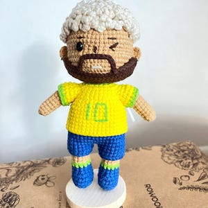 May include: A crochet doll featuring a figure with light skin, a dark brown beard, and curly white hair. The doll wears a bright yellow shirt with the number 10 embroidered in green, blue shorts, and blue shoes with green trim. It stands on a round white base.
