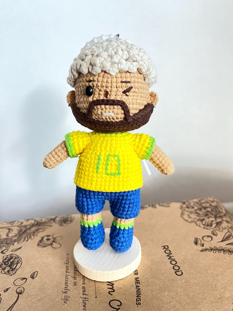 May include: A crochet doll featuring a figure with light skin, a dark brown beard, and curly white hair. The doll wears a bright yellow shirt with the number 10 embroidered in green, blue shorts, and blue shoes with green trim. It stands on a round white base.