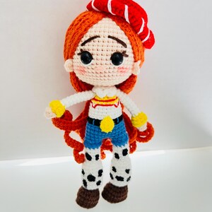 May include: A crocheted doll with red hair, a red and white striped hat, and a blue and white striped shirt. The doll is wearing blue jeans and brown boots with white spots.