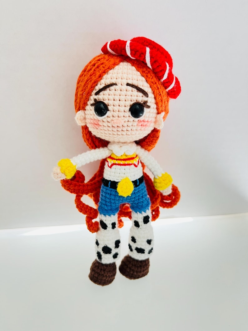 May include: A crocheted doll with red hair, a red and white striped hat, and a blue and white striped shirt. The doll is wearing blue jeans and brown boots with white spots.