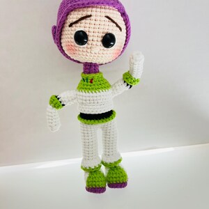 May include: A crocheted toy of Buzz Lightyear from the Toy Story franchise. The toy is wearing a purple helmet, white and green spacesuit, and green boots. The toy has a sad expression on its face.