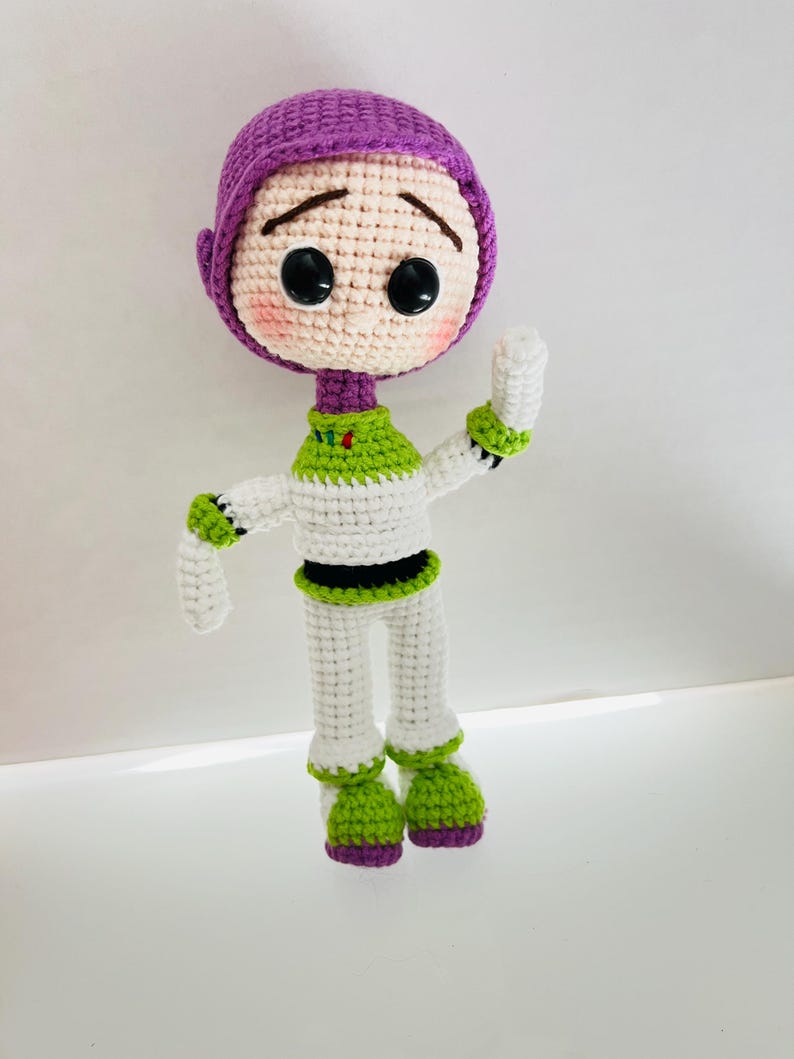 May include: A crocheted toy of Buzz Lightyear from the Toy Story franchise. The toy is wearing a purple helmet, white and green spacesuit, and green boots. The toy has a sad expression on its face.