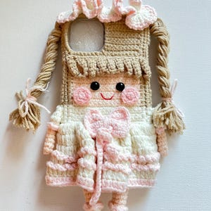 Puede incluir: Una funda de tel&eacute;fono de ganchillo con forma de mu&ntilde;eca con ropa rosa y blanca, cabello casta&ntilde;o y un lazo rosa. La funda tiene una abertura transparente para la c&aacute;mara del tel&eacute;fono.