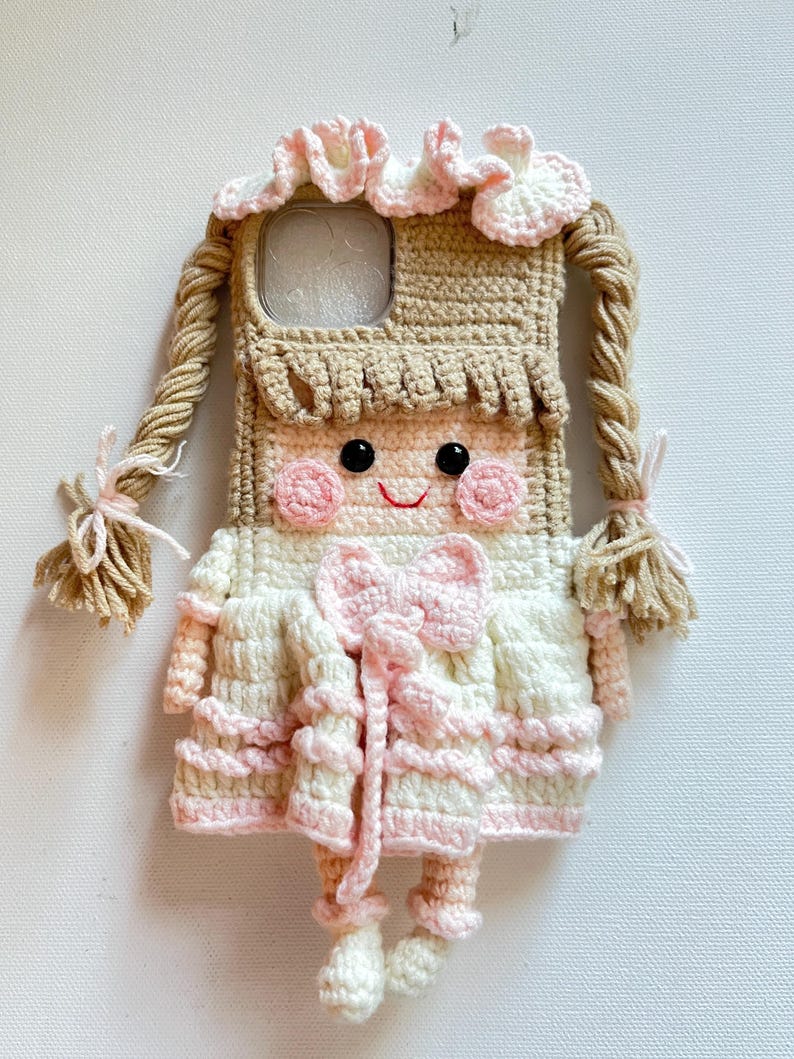 Puede incluir: Una funda de tel&eacute;fono de ganchillo con forma de mu&ntilde;eca con ropa rosa y blanca, cabello casta&ntilde;o y un lazo rosa. La funda tiene una abertura transparente para la c&aacute;mara del tel&eacute;fono.