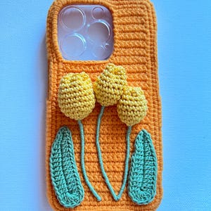 Puede incluir: Funda de tel&eacute;fono de crochet naranja con un dise&ntilde;o de tres flores amarillas y dos hojas verdes.