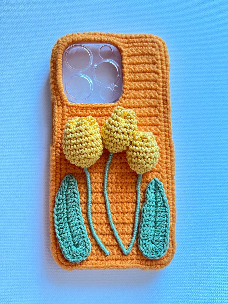 Puede incluir: Funda de tel&eacute;fono de crochet naranja con un dise&ntilde;o de tres flores amarillas y dos hojas verdes.