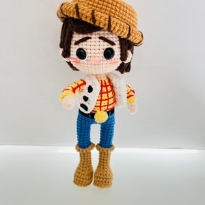 May include: A crocheted toy doll dressed as a cowboy. The doll has brown hair, a brown hat, a red and yellow plaid shirt, blue jeans, and brown boots.