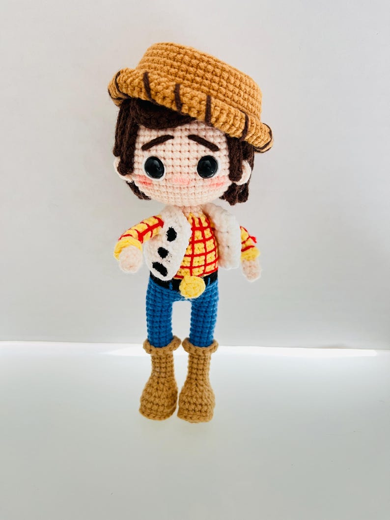 May include: A crocheted toy doll dressed as a cowboy. The doll has brown hair, a brown hat, a red and yellow plaid shirt, blue jeans, and brown boots.