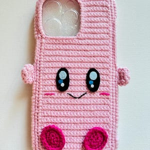 Puede incluir: Funda de tel&eacute;fono de crochet rosa con una cara de dibujos animados, con ojos negros, una peque&ntilde;a sonrisa y mejillas rosadas. La funda tiene un inserto de pl&aacute;stico transparente para la lente de la c&aacute;mara.