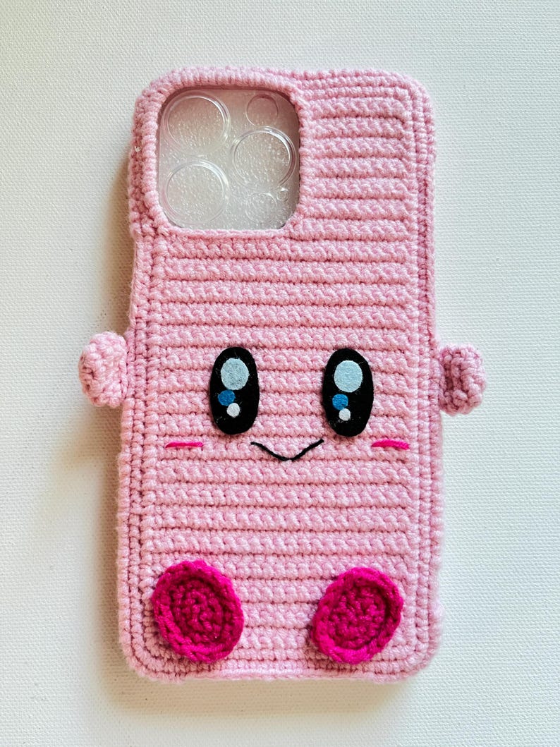 Puede incluir: Funda de tel&eacute;fono de crochet rosa con una cara de dibujos animados, con ojos negros, una peque&ntilde;a sonrisa y mejillas rosadas. La funda tiene un inserto de pl&aacute;stico transparente para la lente de la c&aacute;mara.