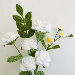 Puede incluir: Un ramo de crochet hecho a mano con rosas blancas y margaritas con centros amarillos. Las flores se colocan sobre tallos y hojas verdes, creando una estética natural y delicada. Un encantador arreglo floral hecho a mano.