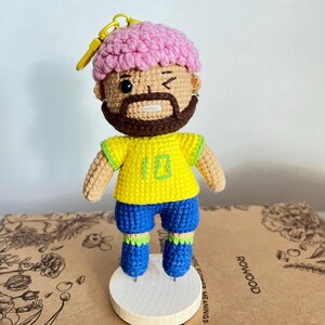 May include: A crocheted doll featuring a figure with pink hair, a brown beard, and a winking expression. The doll wears a yellow jersey with the number 10 in green, blue shorts, and green and blue socks, attached to a yellow plastic keychain ring.