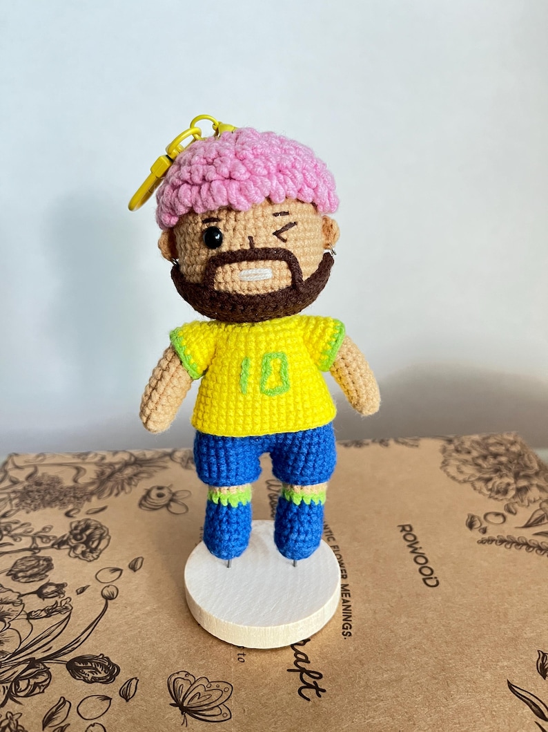 May include: A crocheted doll featuring a figure with pink hair, a brown beard, and a winking expression. The doll wears a yellow jersey with the number 10 in green, blue shorts, and green and blue socks, attached to a yellow plastic keychain ring.