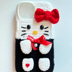 Puede incluir: Funda de tel&eacute;fono Hello Kitty de crochet con un lazo rojo y un dise&ntilde;o blanco y negro. La funda tiene un coraz&oacute;n rojo en la parte inferior.