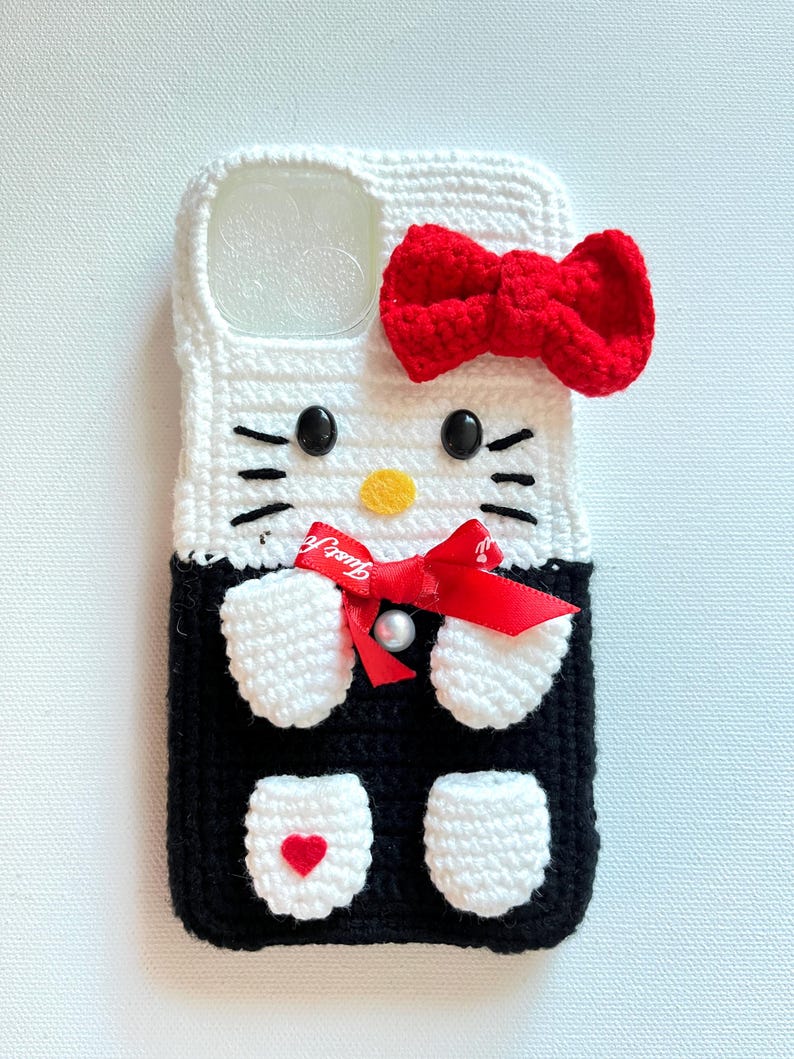 Puede incluir: Funda de tel&eacute;fono Hello Kitty de crochet con un lazo rojo y un dise&ntilde;o blanco y negro. La funda tiene un coraz&oacute;n rojo en la parte inferior.