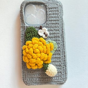 Puede incluir: Una funda de tel&eacute;fono de crochet gris con una flor amarilla y una flor blanca como adorno.