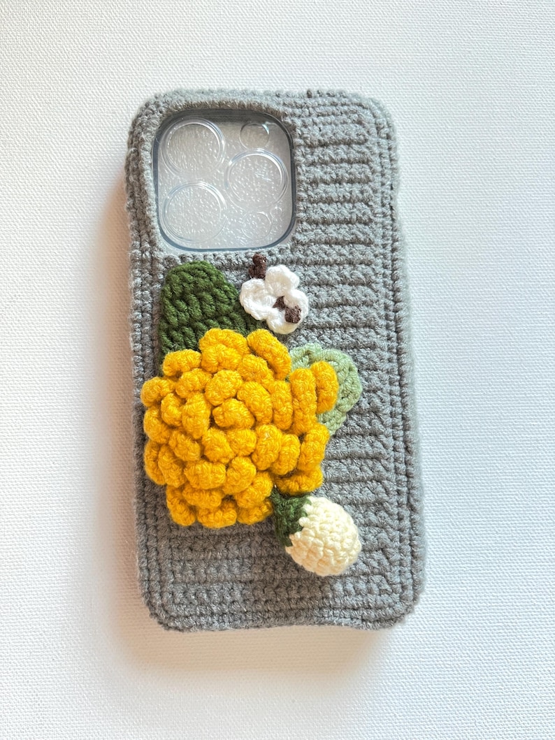Puede incluir: Una funda de tel&eacute;fono de crochet gris con una flor amarilla y una flor blanca como adorno.