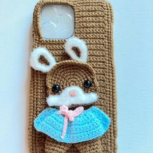 Puede incluir: Una funda de tel&eacute;fono de crochet marr&oacute;n con un conejito blanco sujeto a la parte delantera. El conejito lleva un vestido azul y rosa y orejas blancas.