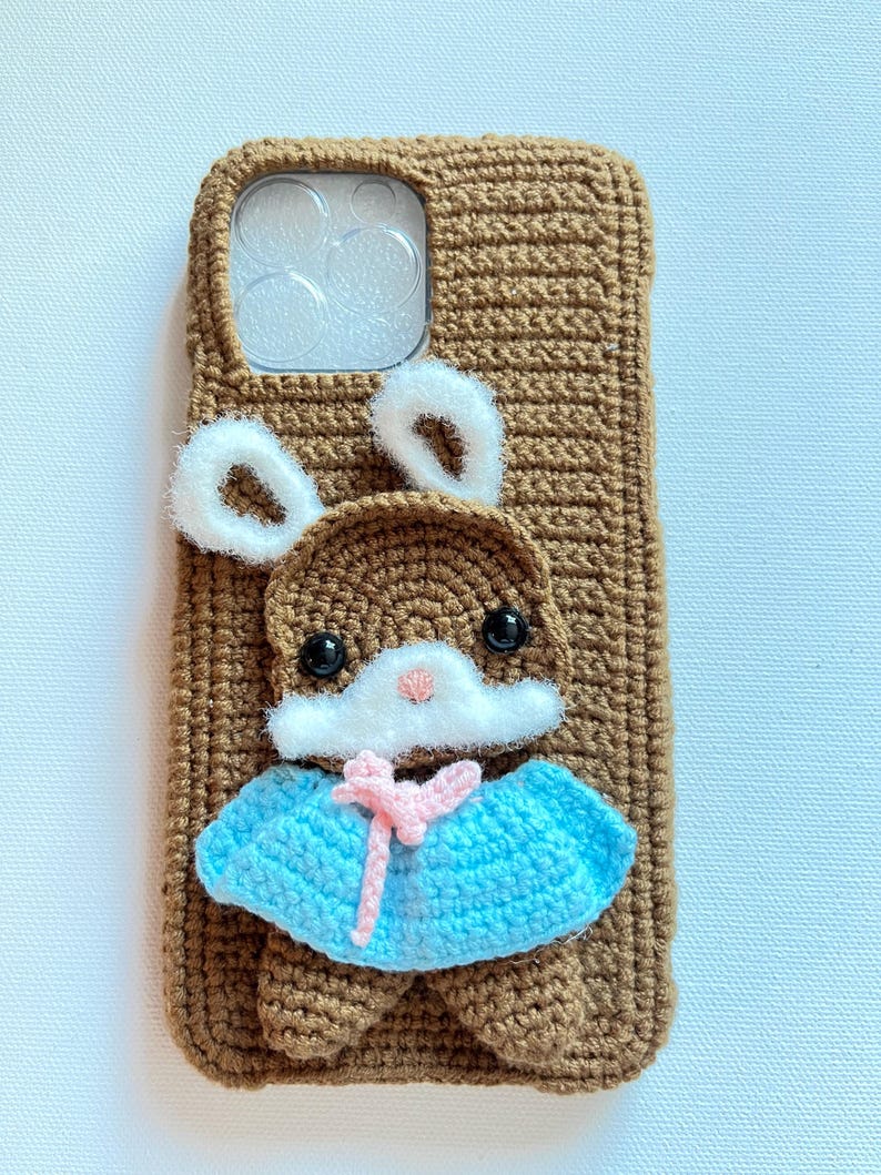 Puede incluir: Una funda de tel&eacute;fono de crochet marr&oacute;n con un conejito blanco sujeto a la parte delantera. El conejito lleva un vestido azul y rosa y orejas blancas.