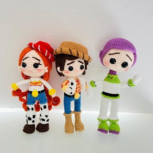 May include: Three crocheted plush toys inspired by the characters from the Toy Story movie franchise. The toys are Woody, Jessie, and Buzz Lightyear. Woody is wearing a brown cowboy hat, a red and white plaid shirt, blue jeans, and brown boots. Jessie is wearing a red and white hat, a yellow shirt, blue jeans, and brown boots. Buzz Lightyear is wearing a purple helmet, a green shirt, white pants, and green boots.