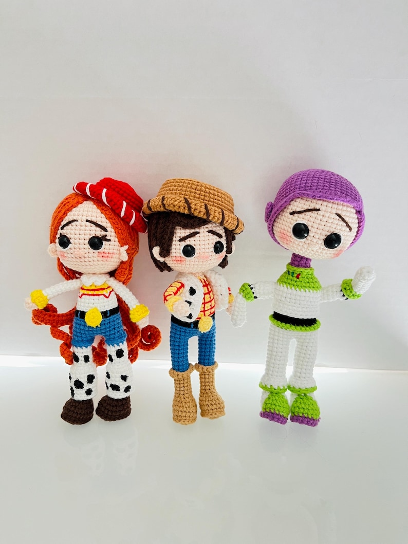 May include: Three crocheted plush toys inspired by the characters from the Toy Story movie franchise. The toys are Woody, Jessie, and Buzz Lightyear. Woody is wearing a brown cowboy hat, a red and white plaid shirt, blue jeans, and brown boots. Jessie is wearing a red and white hat, a yellow shirt, blue jeans, and brown boots. Buzz Lightyear is wearing a purple helmet, a green shirt, white pants, and green boots.