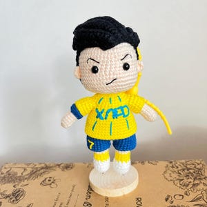 K&ouml;nnte beinhalten: Eine geh&auml;kelte Puppe mit schwarzem Haar, einem gelben Trikot mit blauem Text und Akzenten, blauer Hose mit der gelben Nummer 7 und wei&szlig;en Schuhen. Die Puppe steht auf einem runden Holzsockel und hat eine gelbe Kordel am R&uuml;cken befestigt.