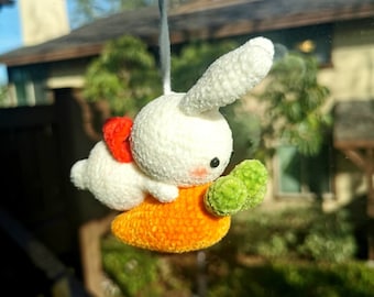 Pode incluir: Um coelho branco de crochê sentado em uma cenoura laranja de crochê com folhas verdes. O coelho tem um laço vermelho na orelha.
