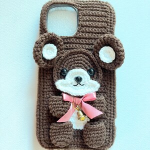 Puede incluir: Una funda de tel&eacute;fono de crochet marr&oacute;n con un dise&ntilde;o de oso de peluche. El oso tiene orejas blancas, una cara blanca y un lazo rosa. La funda tiene un recorte transparente para la c&aacute;mara del tel&eacute;fono.