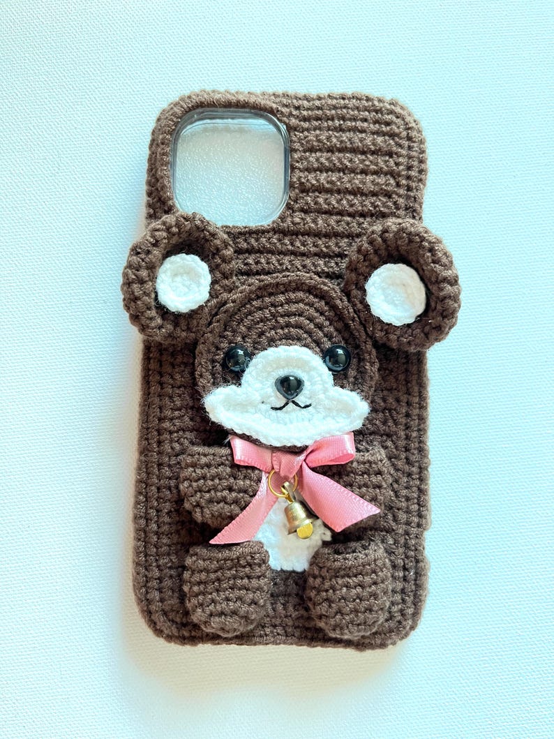 Puede incluir: Una funda de tel&eacute;fono de crochet marr&oacute;n con un dise&ntilde;o de oso de peluche. El oso tiene orejas blancas, una cara blanca y un lazo rosa. La funda tiene un recorte transparente para la c&aacute;mara del tel&eacute;fono.