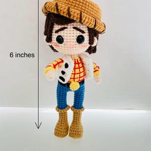 May include: A crocheted toy figure of a cowboy, standing 6 inches tall. The figure is wearing a brown hat, a red and yellow plaid shirt, blue jeans, and brown boots. The figure has brown hair and a friendly face.